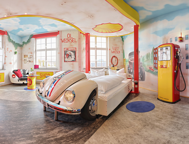 Das Bild zeigt ein thematisches Hotelzimmer im Retro-Stil. Im Zentrum steht ein Bett, das wie ein sportlicher VW Käfer gestaltet ist. Die Wände sind mit bunten Wandmalereien, darunter eine alte Tankstelle, dekoriert. Das Ambiente erinnert an eine Autowerkstatt der Vergangenheit.