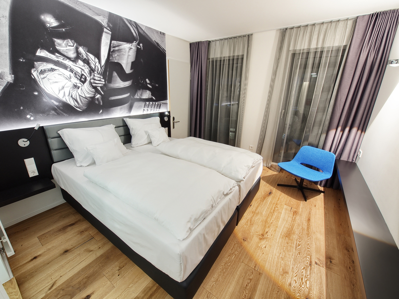 Modern eingerichtetes Hotelzimmer mit komfortablem Doppelbett, blauem Sessel in der Ecke sowie Bild an der Wand.
