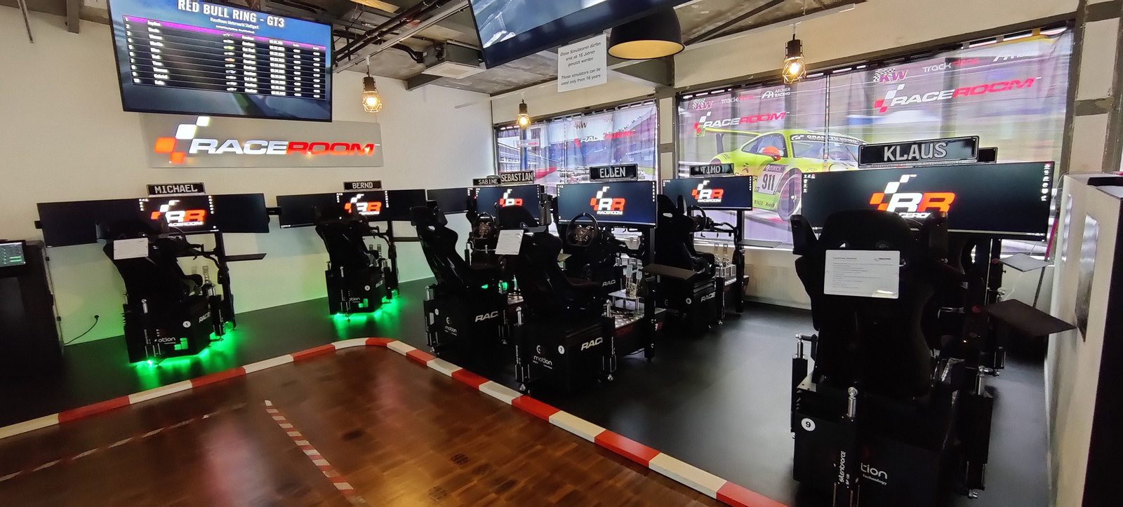 In einem Raum stehen mehrere Rennsimulationseinheiten mit Bildschirmen und Rennsitzen. Jede Einheit hat einen Namen über dem Bildschirm. Im Hintergrund ist ein großer Monitor, der Ergebnisse des Red Bull Ring - GT3 anzeigt. Die Umgebung ist modern und gut beleuchtet.