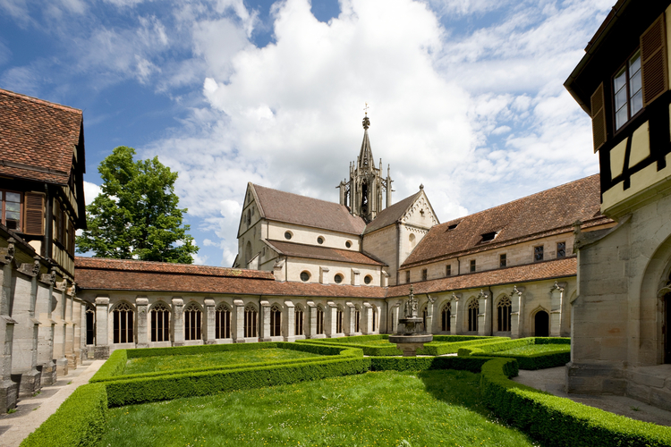Eine grüne Klostergartenlandschaft mit prächtigen Hecken und einem zentralen Brunnen, umgeben von historischen Gebäuden mit Fachwerk und gotischen Fenstern. Im Hintergrund ragt eine Kirche mit einem hohen, spitzen Turm und hellem Himmel.