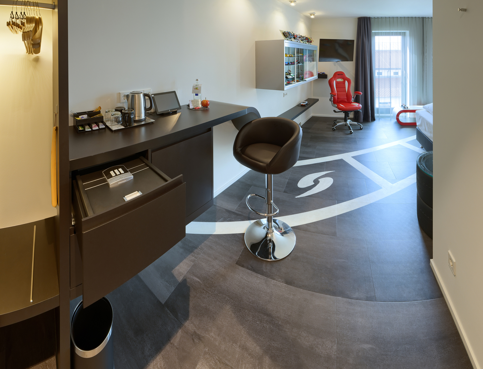 Das Bild zeigt ein modernes Hotelzimmer mit einem minimalistischen Design. Links ist ein Schreibtisch mit verschiedenen Annehmlichkeiten wie einem Wasserkocher. Rechts steht ein roter Bürostuhl und im Hintergrund ein Bett. Der Boden hat ein markantes, kurviges Muster.