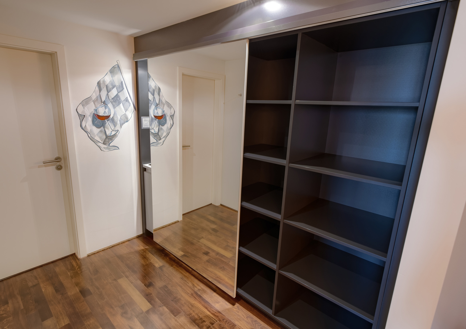 Ein Innenraum zeigt einen schwarzen Regalschrank mit mehreren offenen Fächern. Gegenüber befindet sich ein Spiegel, der die Wand reflektiert, auf der eine dekorative Flagge abgebildet ist. Der Boden besteht aus Holz und verleiht dem Raum eine warme Atmosphäre.