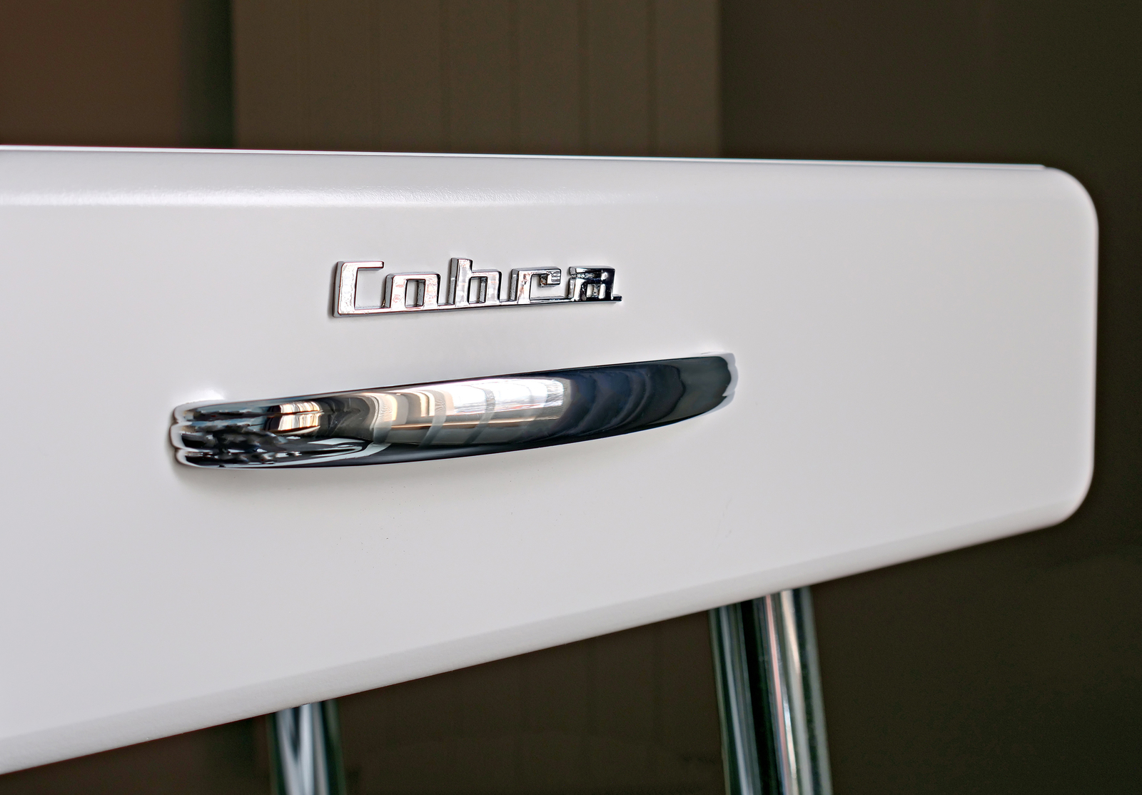 Ein naher Blick auf eine weiße Kühlschranktür mit einem glänzenden Griff. Oben ist das Logo "Cobra" in metallischer Schrift angebracht. Der Griff hat eine geschwungene Form und reflektiert das Licht, während die Tür selbst glatt und modern wirkt.