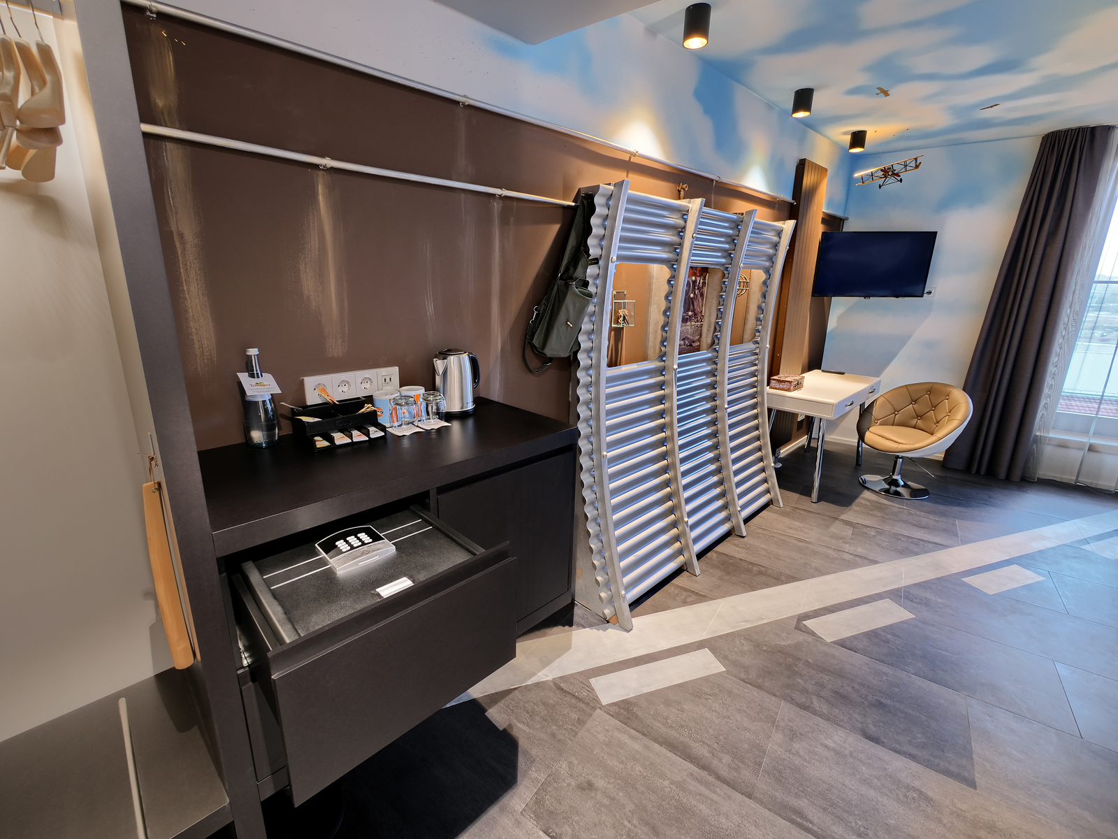 Ein modernes Hotelzimmer mit einem Schreibtisch, einem Fernseher an der Wand und einem stilvollen Stuhl. Neben einem Kühlschrank stehen Wasserkocher und Gläser auf einem schwarzen Tisch. Der Raum hat eine helle Farbgestaltung mit Wolkenmotiven an der Decke.