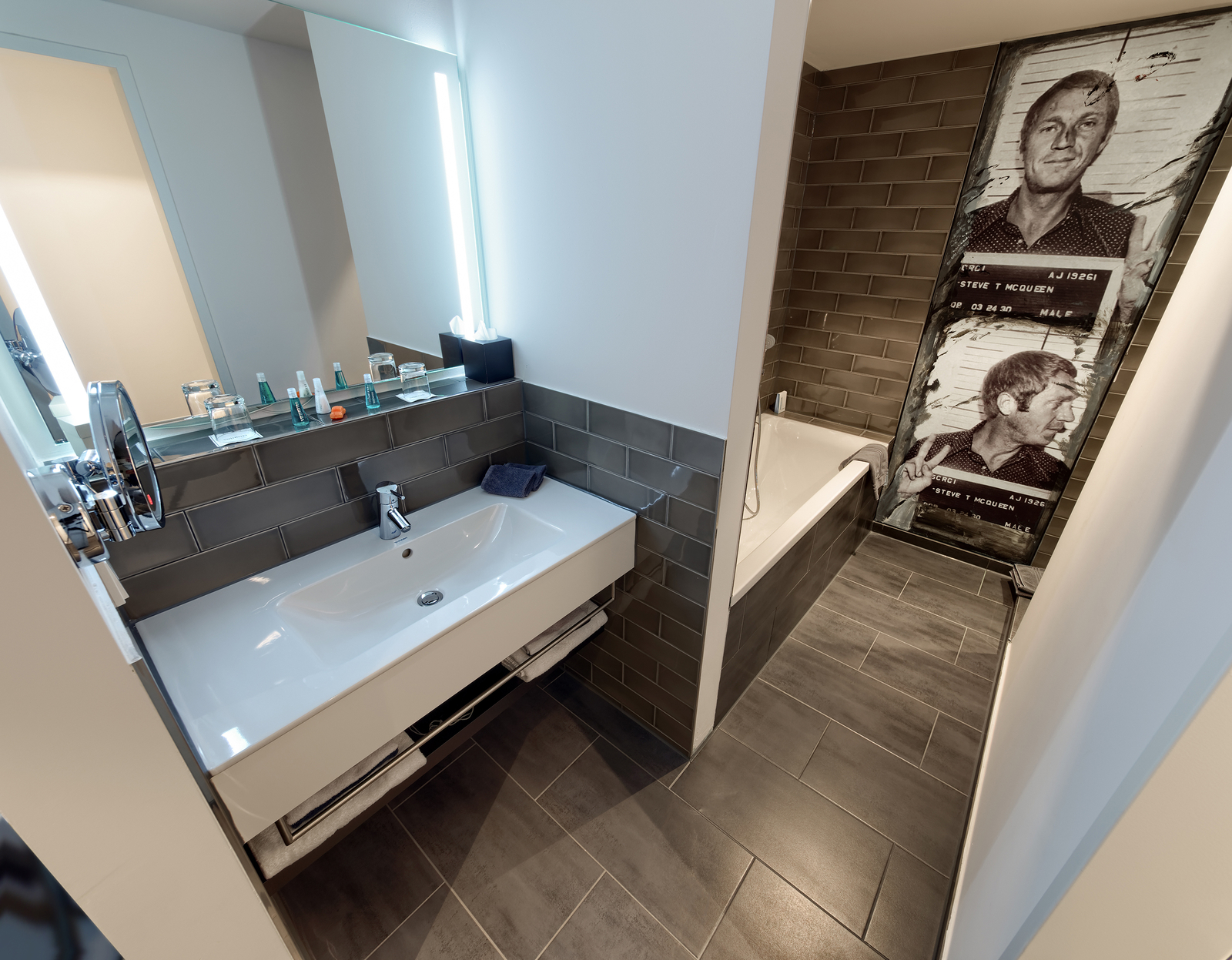 Ein modernes Badezimmer mit einem weißen Waschbecken und einem großen Wandspiegel. Auf der Ablage stehen verschiedene Pflegeprodukte. Im Hintergrund sieht man eine Badewanne und eine Wand mit einem auffälligen Schwarz-Weiß-Bild einer Person. Der Boden ist mit grauen Fliesen gestaltet.