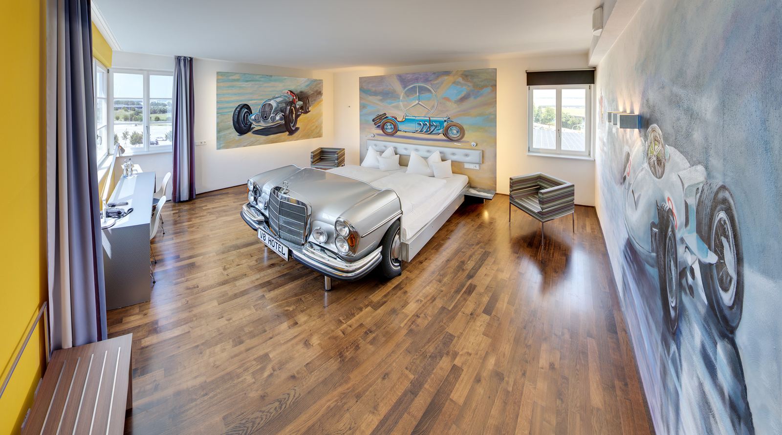 Das Bild zeigt ein stilvolles Hotelzimmer mit einem einzigartigen Bett, das wie ein silberner Oldtimer aussieht. Die Wände sind mit großformatigen Automobilmotiven dekoriert, und der Boden besteht aus Holz. Große Fenster lassen Tageslicht herein und bieten einen Blick nach draußen.