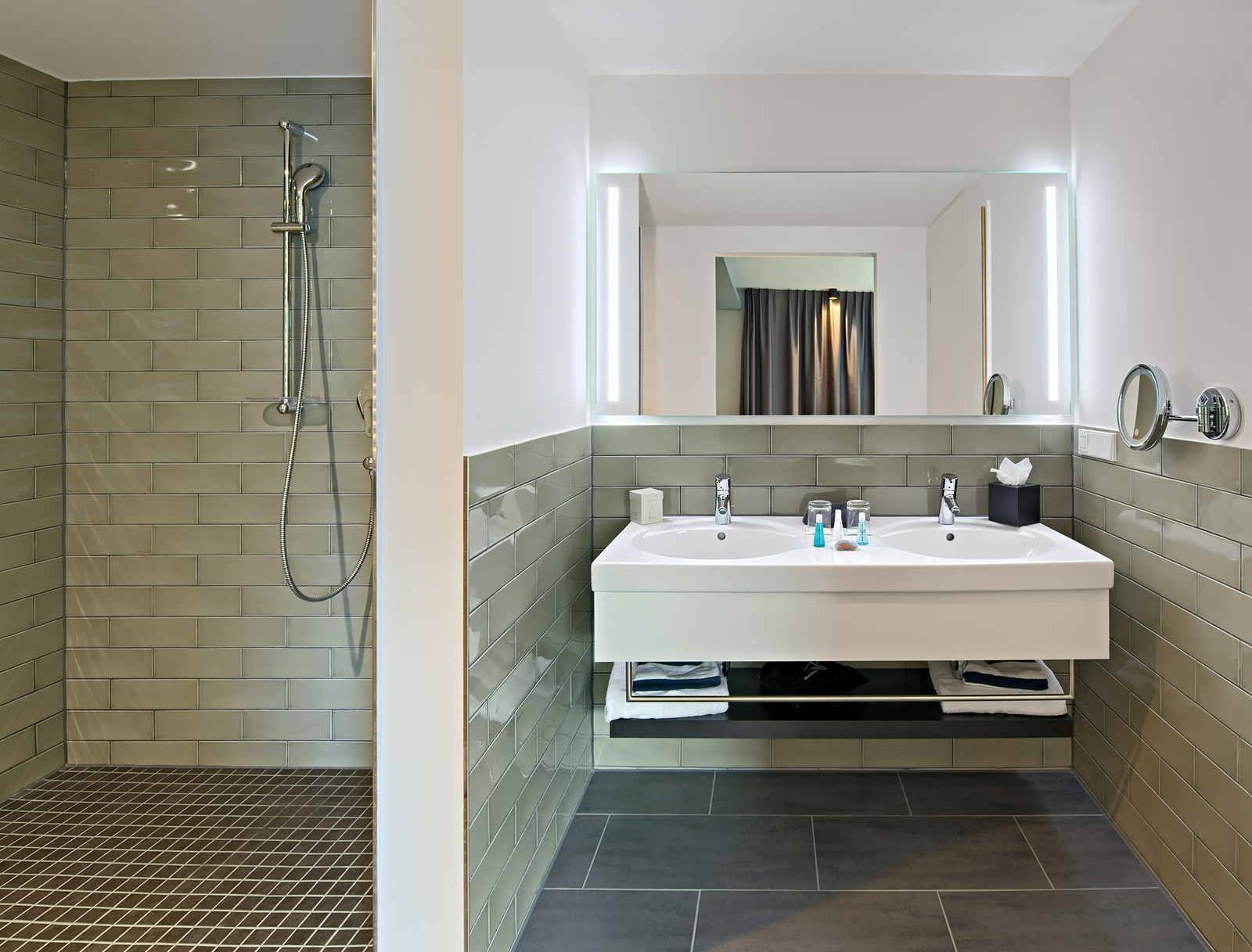Das Bild zeigt ein modernes Badezimmer mit einer Dusche in der linken Ecke und einem Doppelwaschbecken an einer Wand. Der Waschbereich ist mit zwei Wasserhähnen und flachen Regalen ausgestattet. Die Wände sind in neutralen Farben gehalten, und es gibt ein Spiegel über dem Waschbecken.