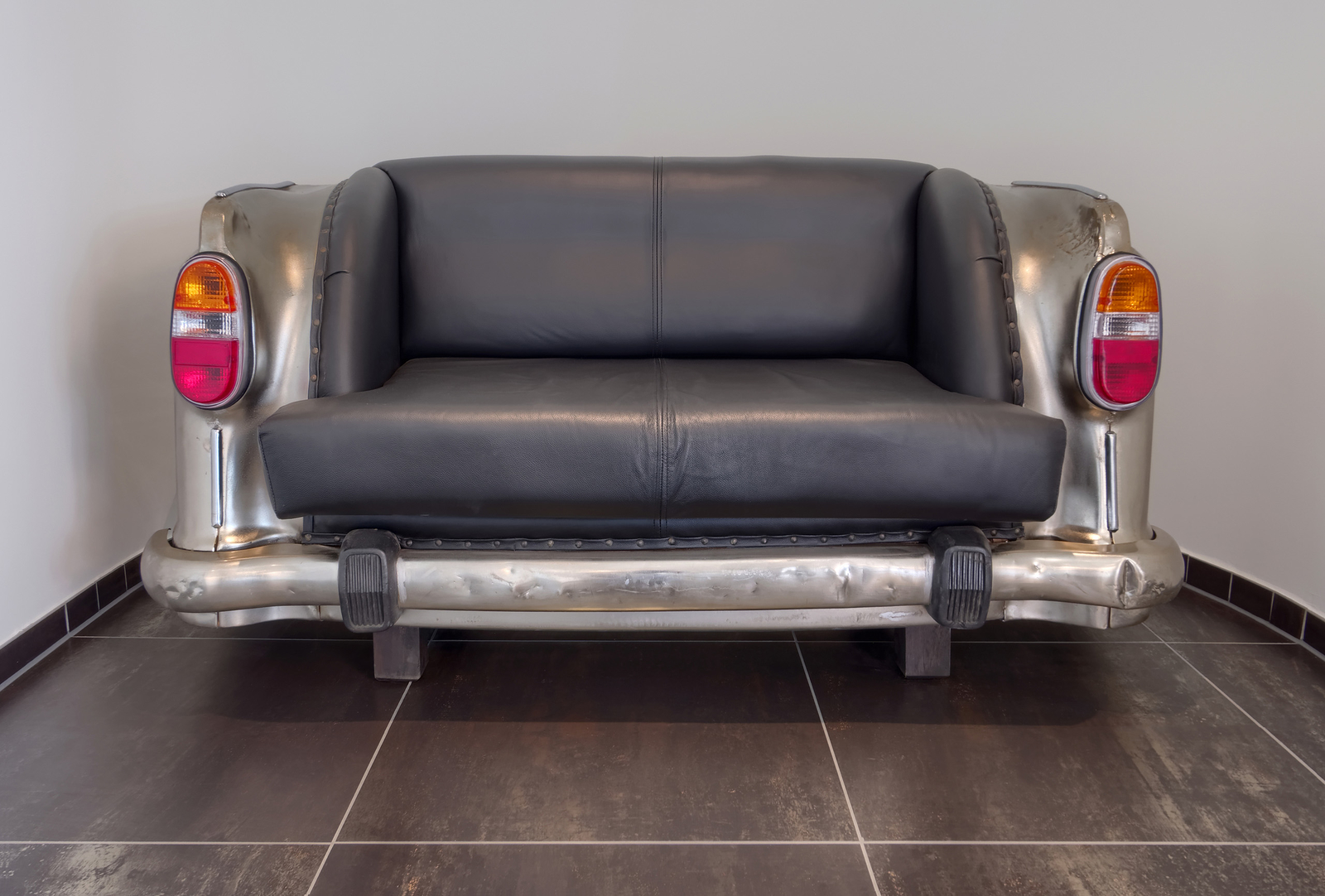 Ein schwarzes Sofa, das auf der Rückseite einem Autodesign ähnelt. Die Armlehnen sind rund und metallisch, während die Sitze aus schwarzem Leder bestehen. Das Sofa steht auf einem gefliesten Boden und vermittelt einen modernen, kreativen Stil.