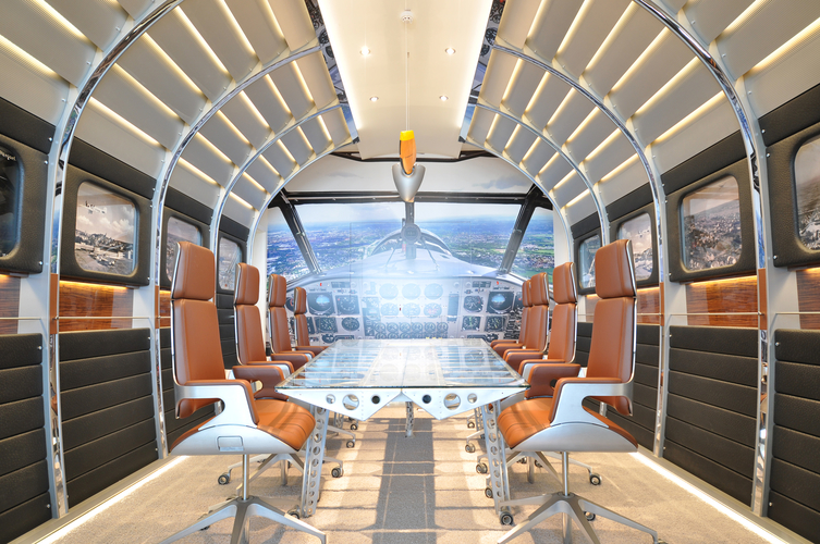 Ein moderner Besprechungsraum im Stil eines Flugzeugcockpits. Er ist mit braunen Drehstühlen und einem gläsernen Tisch ausgestattet. An den Wänden sind Bilder von Lanzplätzen zu sehen, und in der Mitte befindet sich ein großes Fenster mit Blick auf eine Landschaft.
