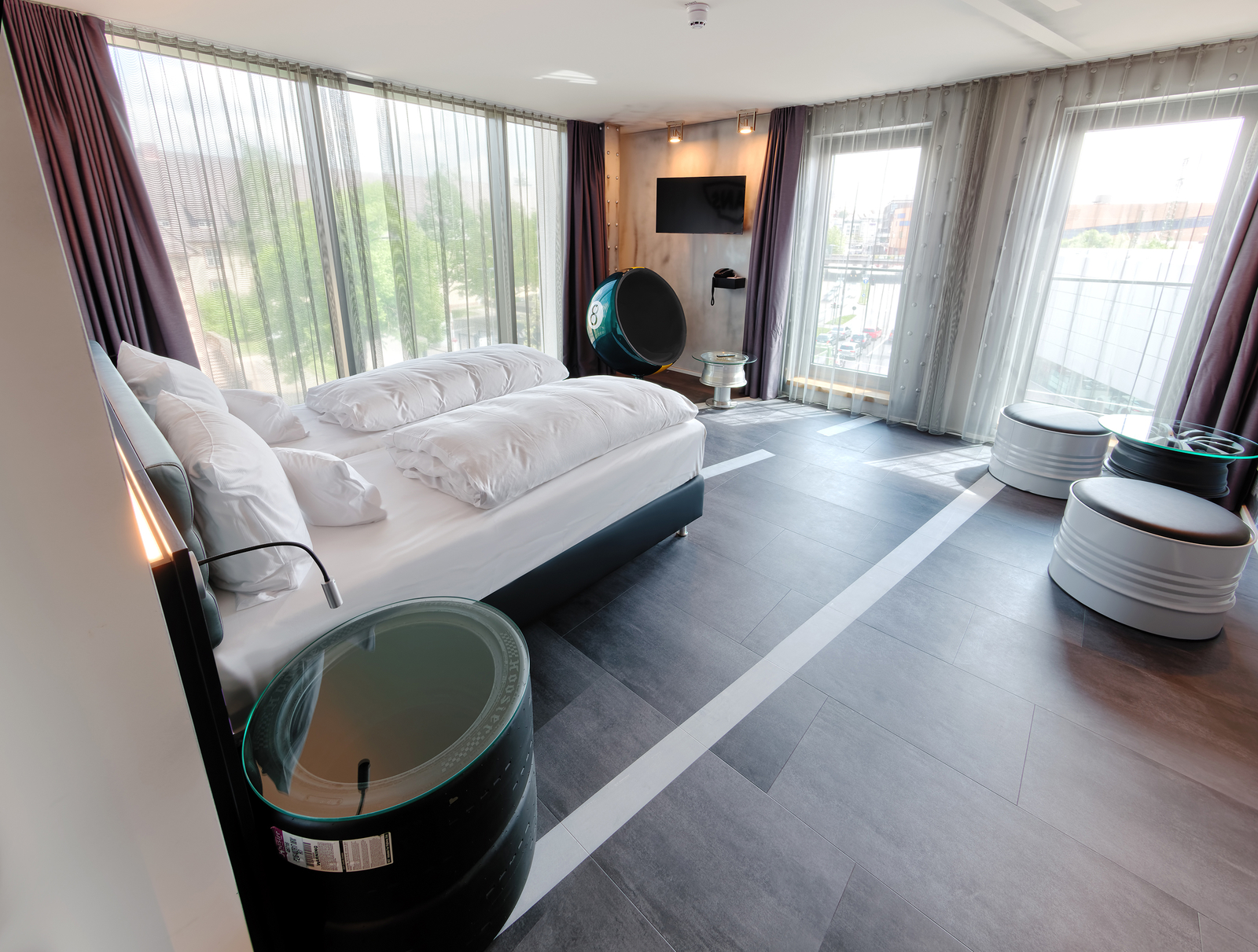 Das Bild zeigt ein modernes Hotelzimmer mit einem großen Doppelbett, umgeben von Fensterfronten. Es gibt runde Tische aus Reifen sowie einen Fernseher an der Wand. Tageslicht strömt durch die Vorhänge und schafft eine einladende Atmosphäre.