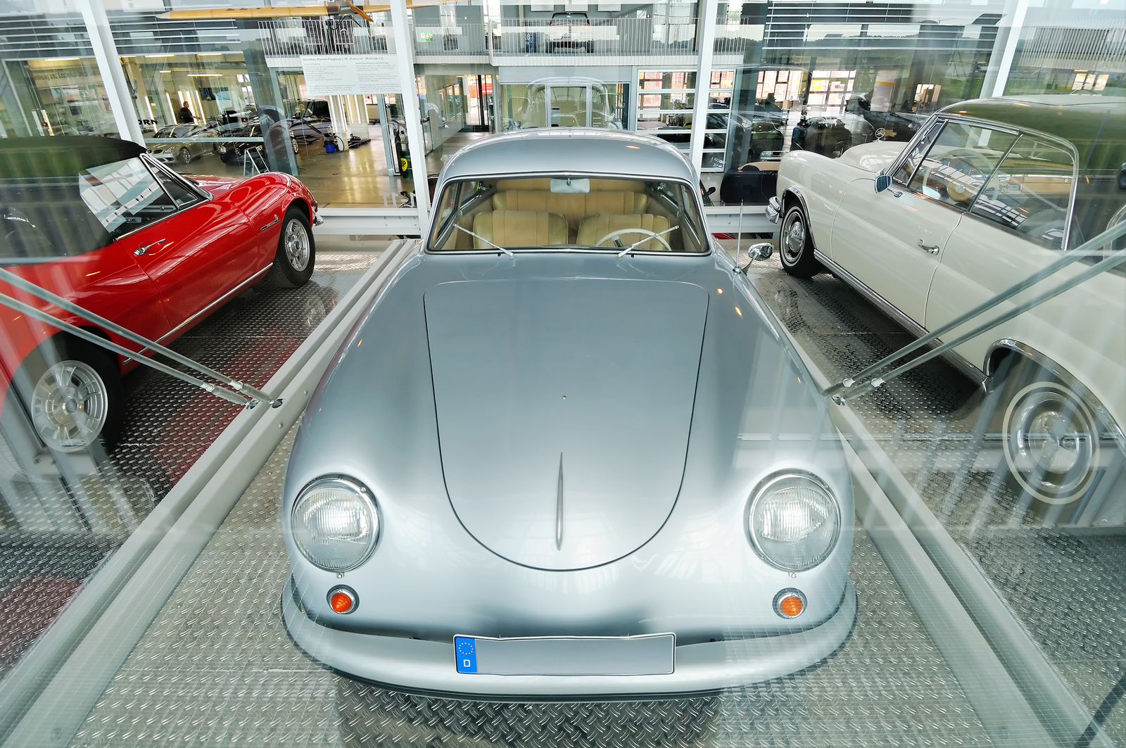 Ein silbernes Oldtimer-Auto steht zentral in einem modernen Ausstellungsraum in der Motorworld. Der Wagen hat runde Scheinwerfer und eine elegante Form. Zwei weitere Autos, ein rotes und ein weißes, sind auf der linken und rechten Seite des silbernen Fahrzeugs sichtbar.