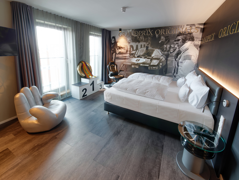 Grandprix-Themenzimmer im V8 Hotel eingerichtet mit einem gemütlichen Doppelbett gegenüber eines Siegertreppchens und einem Sessel in Form einer Hand