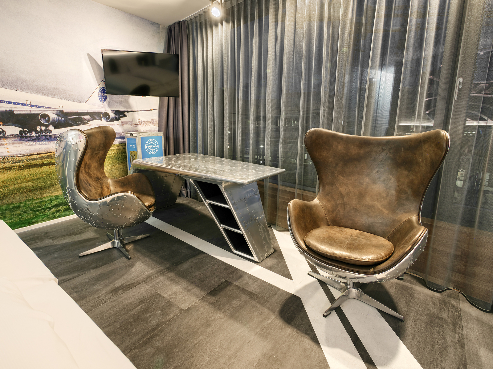 Ein modernes Zimmer mit einem schlichten Schreibtisch aus Metall und zwei stilvollen Sesseln mit einer Kombination aus Edelstahl und Leder. Der Raum hat eine große Fensterfront mit Vorhängen und einem großen Fernseher an der Wand.