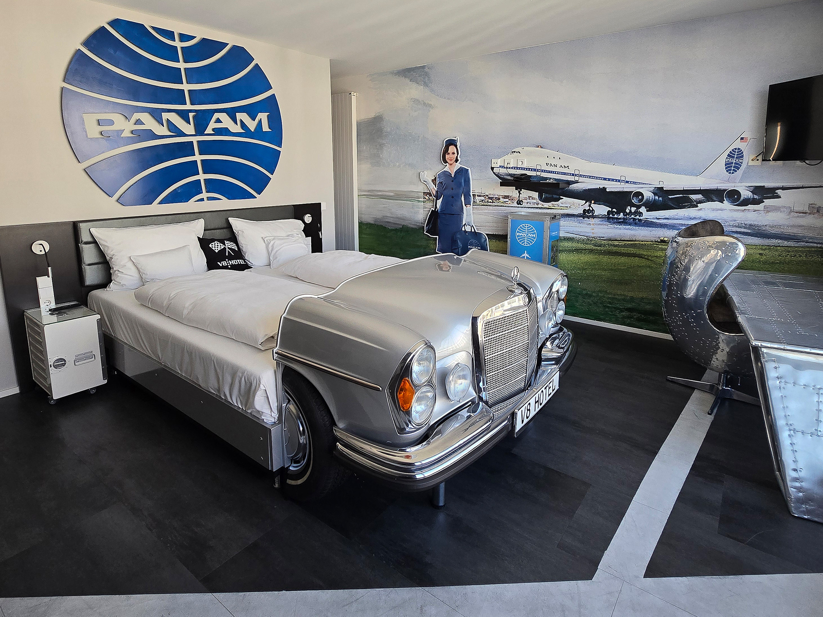 Ein Hotelzimmer im Vintage-Stil mit einem Bett, das wie ein silberner Oldtimer aussieht. An der Wand ist das Logo von Pan Am zu sehen, zusammen mit einem Wandbild eines Flugzeugs und einer Stewardess in Uniform. Ein futuristischer Schreibtisch ergänzt die Einrichtung.