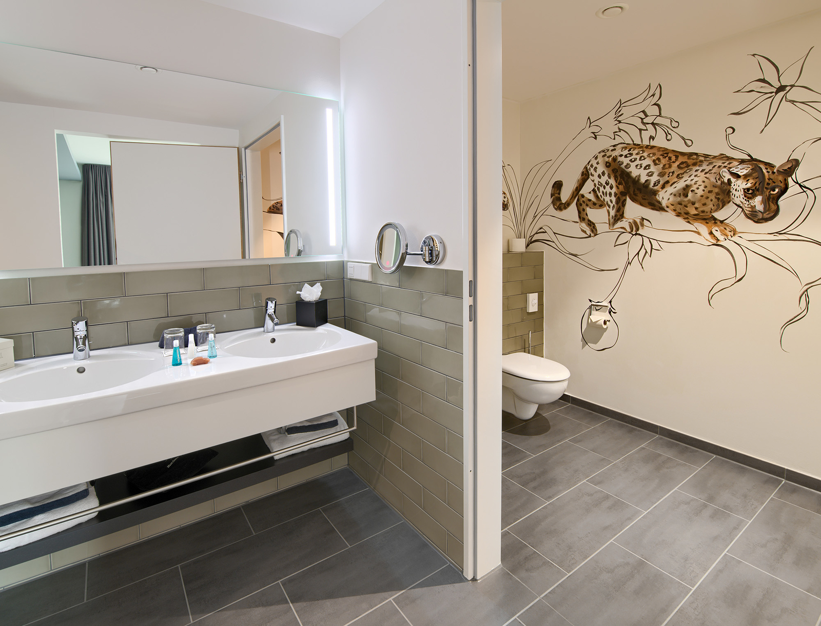 Modernes Badezimmer mit Doppelwaschbecken und Wandspiegel. An der Wand ist ein kunstvolles Leopard-Motiv zu sehen. Der Boden ist mit grauen Fliesen ausgelegt, und es gibt eine separate Toilette im Hintergrund. Hygieneartikel sind auf dem Waschbecken platziert.