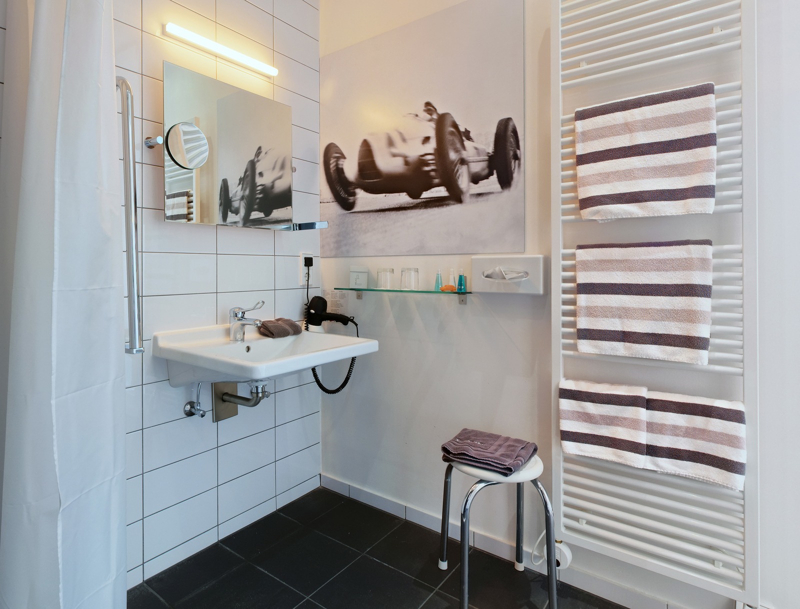 Das Bild zeigt ein modernes, barrierefreies Badezimmer mit einem Waschbecken, einem Spiegel und einem Handtuchhalter. An der Wand ist ein großes schwarz-weißes Foto eines Rennwagens. Es gibt einen Stuhl und unter dem Waschbecken ein Handtuch. Die Wände sind gefliest und hell.