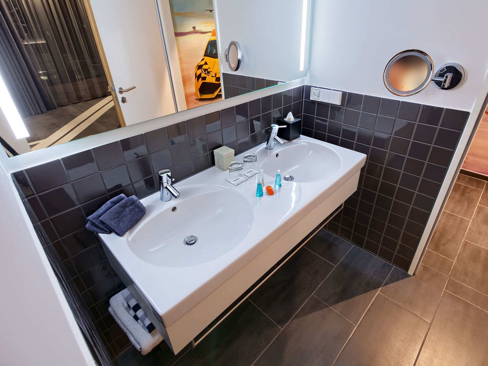 Das Bild zeigt ein modernes Badezimmer mit einem doppelten Waschbecken. Auf dem Becken sind verschiedene Pflegeprodukte und zwei Handtücher platziert. Der Boden ist gefliest, und die Wände sind in dunklem Grau gefliest, was der Raumgestaltung ein elegantes Aussehen verleiht.