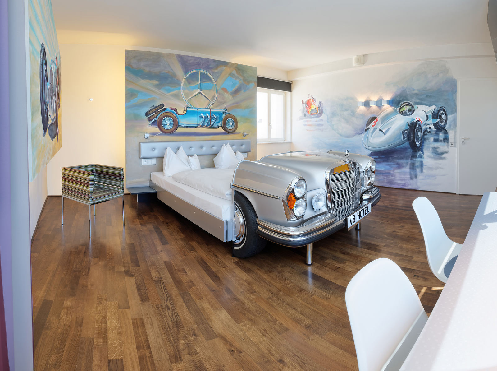 Graues Mercedes-Bett an einer bemalten Wand, die einen Oldtimer von Mercedes zeigt eggenüber eines Schreibtischs mit zwei weißen Stühlen