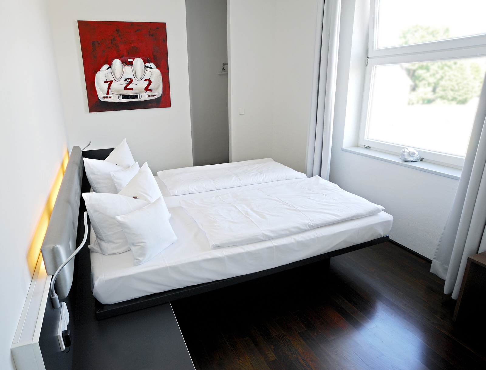 Das Bild zeigt ein modernes Schlafzimmer mit zwei getrennten Betten, die mit weißen Bettwäsche und Kissen dekoriert sind. An der Wand hängt ein Kunstwerk mit der Aufschrift "722". Ein großes Fenster mit Vorhängen lässt Tageslicht herein. Der Boden ist dunkelholzfarben.