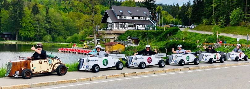 Sechs Personen fahren in kleinen, bunten Autos entlang einer Straße am Ufer eines Sees. Alle tragen Helme und lächeln, während sie Daumen nach oben zeigen. Im Hintergrund ist ein großes, traditionelles Gebäude und eine grüne Landschaft sichtbar.