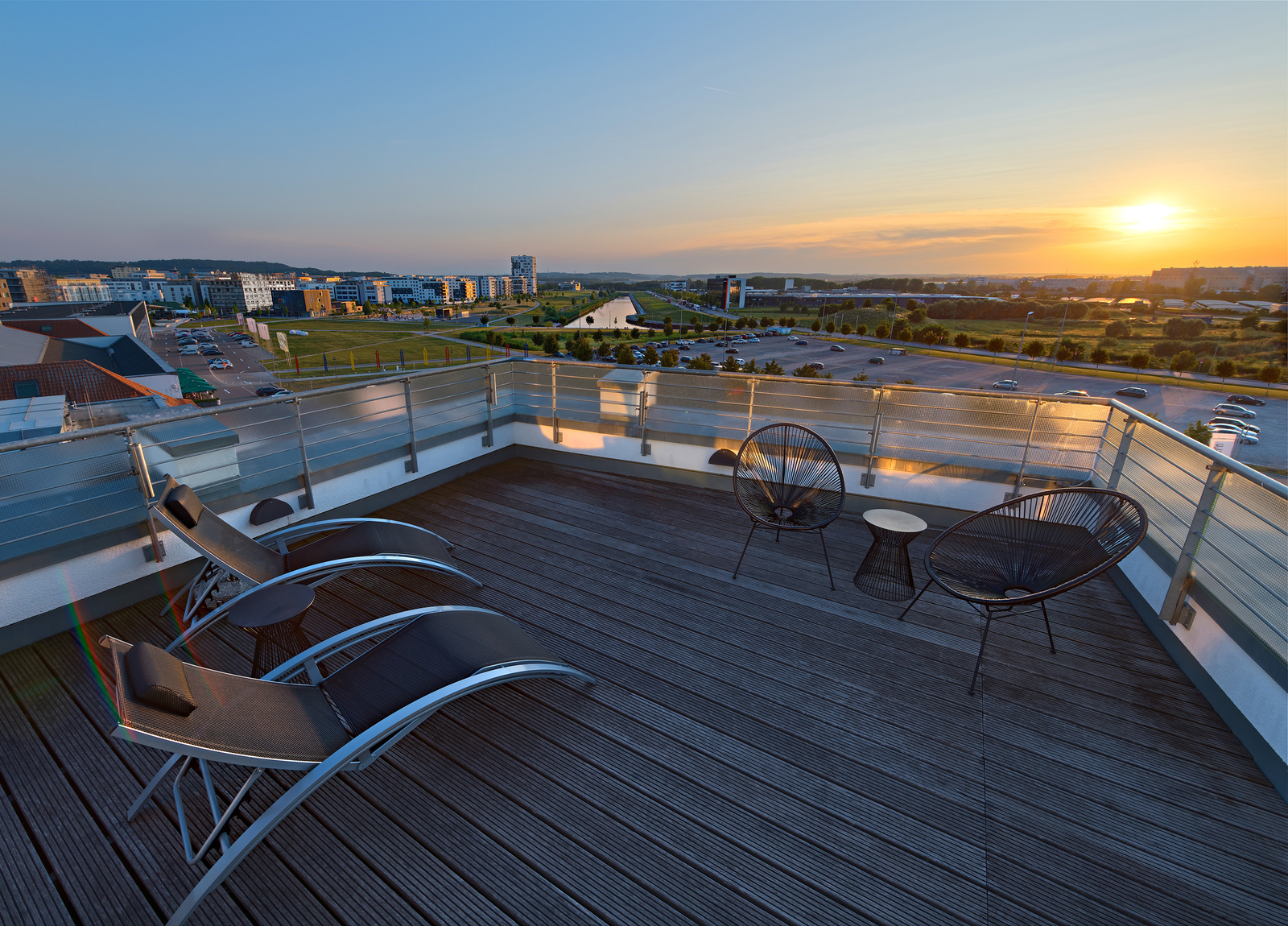 Eine Dachterrasse mit zwei modernen Liegen und einem kleinen Tisch. Im Hintergrund ist ein weitläufiger Ausblick auf eine Stadtlandschaft, während die Sonne am Horizont untergeht und den Himmel in warmen Farbtönen erleuchtet.