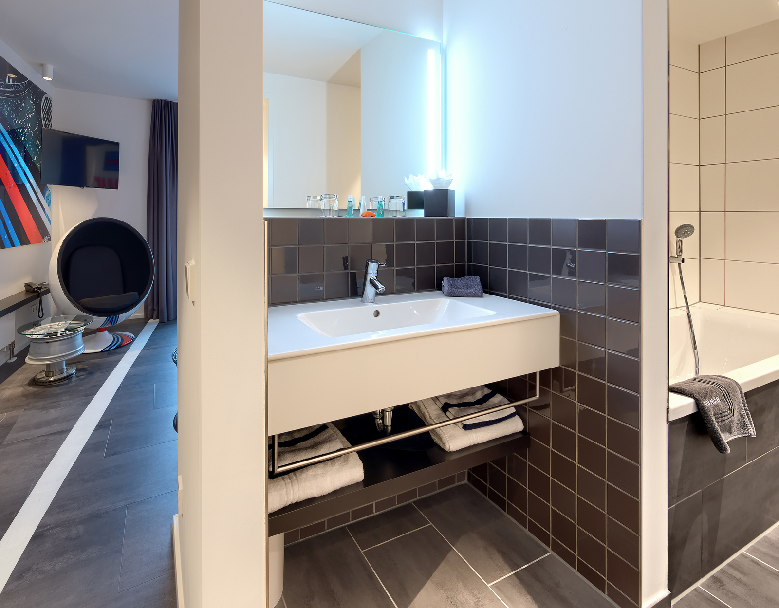 Ein modernes Badezimmer mit einem weißen Waschbecken und einem großen Spiegel, umgeben von dunklen Fliesen. Unter dem Waschbecken sind gerollte Handtücher verstaut. Auf der rechten Seite befindet sich eine Badewanne. Im Hintergrund ist ein stilvolles Wohnzimmer mit einem runden Sessel und einem Fernseher sichtbar.