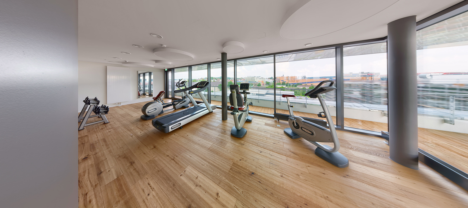 Ein modernes Fitnessstudio mit großen Fenstern, die Blick auf die Umgebung bieten. Es sind mehrere Trainingsgeräte wie Laufband, Fahrräder und Crosstrainer in einem hellen Raum mit Holzboden angeordnet. Die Atmosphäre wirkt einladend und freundlich.