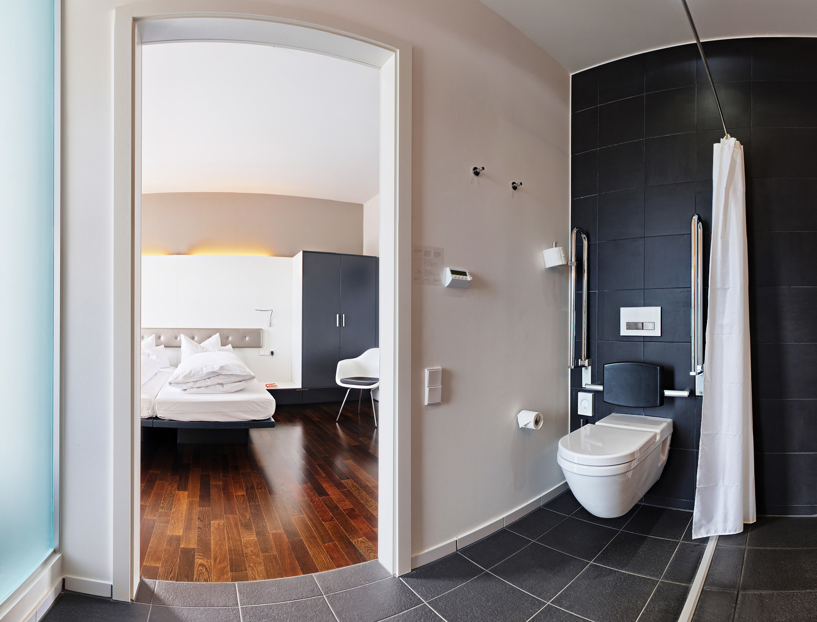 Ein modernes, barrierefreies Badezimmer mit einem weißen WC, einer Dusche mit Vorhang und Haltegriffen. Im Hintergrund ist ein angrenzendes Schlafzimmer sichtbar, das mit einem großen Bett und einem Schrank ausgestattet ist. Der Boden besteht aus dunklem Holz, und die Wände sind hell gestaltet.