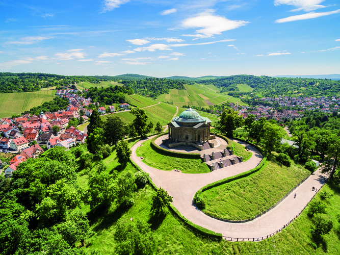 Eine Panoramaansicht zeigt ein historisches Gebäude auf einem Hügel, umgeben von üppigem Grün und Weinbergen. Im Hintergrund ist ein malerisches Dorf mit roten Dächern zu sehen. Der Himmel ist klar und sonnig, was eine einladende Atmosphäre schafft.