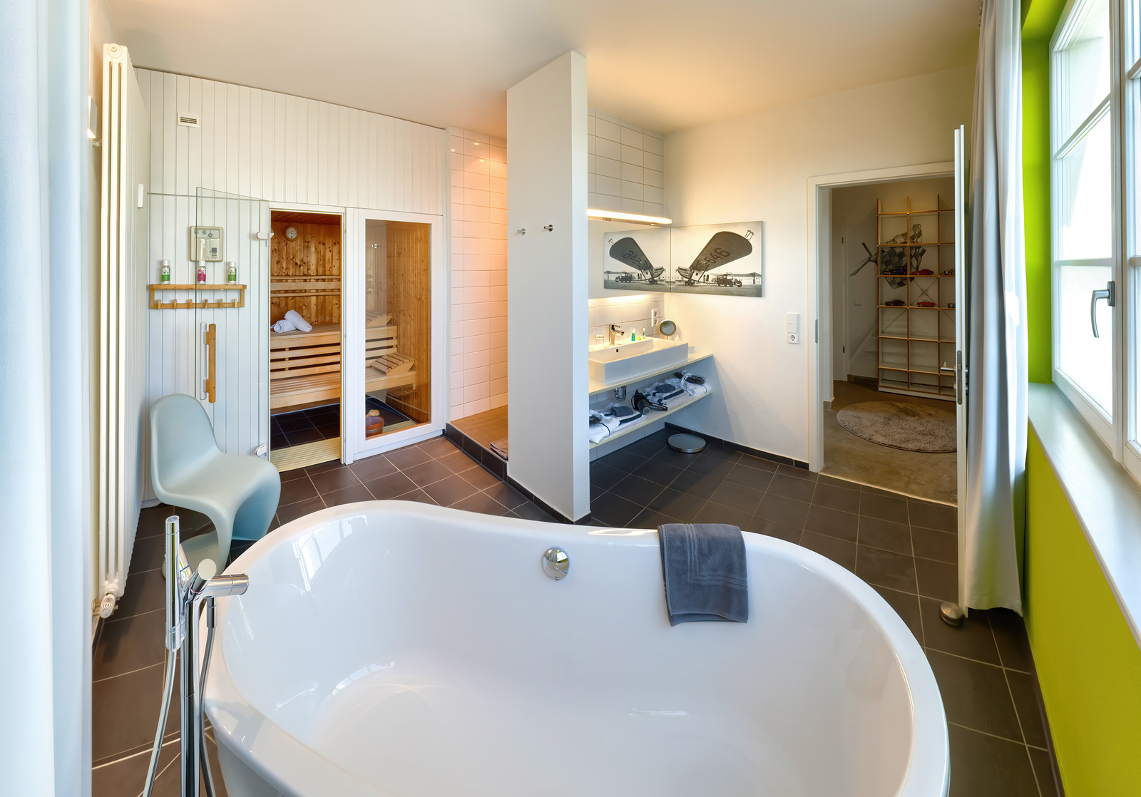 Ein modernes Badezimmer mit einer freistehenden Badewanne und einer Sauna. Der Raum ist hell und stilvoll eingerichtet, mit einem Waschbecken und einem Regal. Große Fenster lassen Tageslicht herein und erzeugen eine entspannende Atmosphäre.