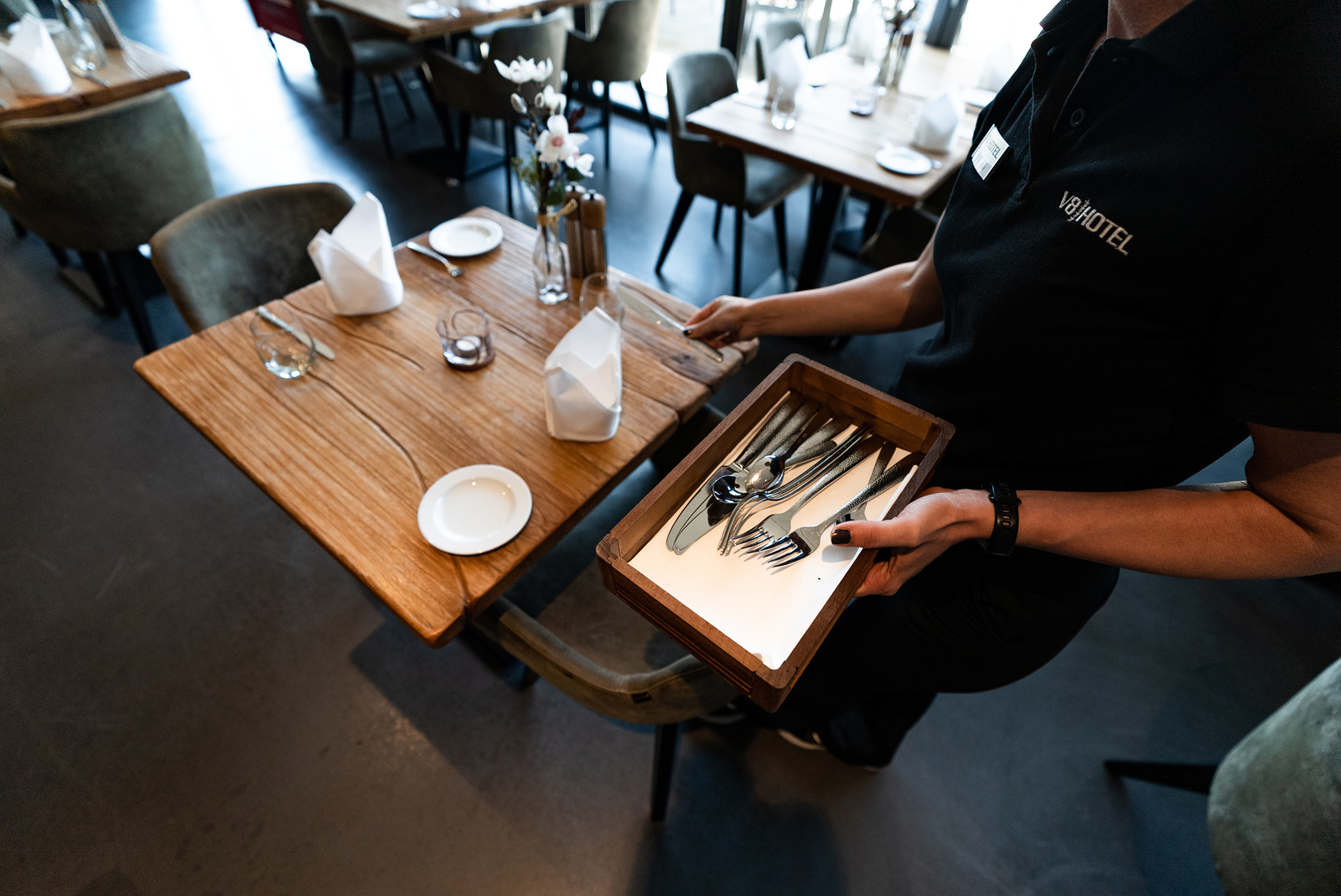 Eine Kellnerin trägt eine Holzschachtel mit Besteck, darunter Gabeln, Messer und Löffel. Der Tisch ist gemütlich gedeckt mit Tellern, Gläsern und einer kleinen Blumenvase. Das Ambiente wirkt modern und einladend.