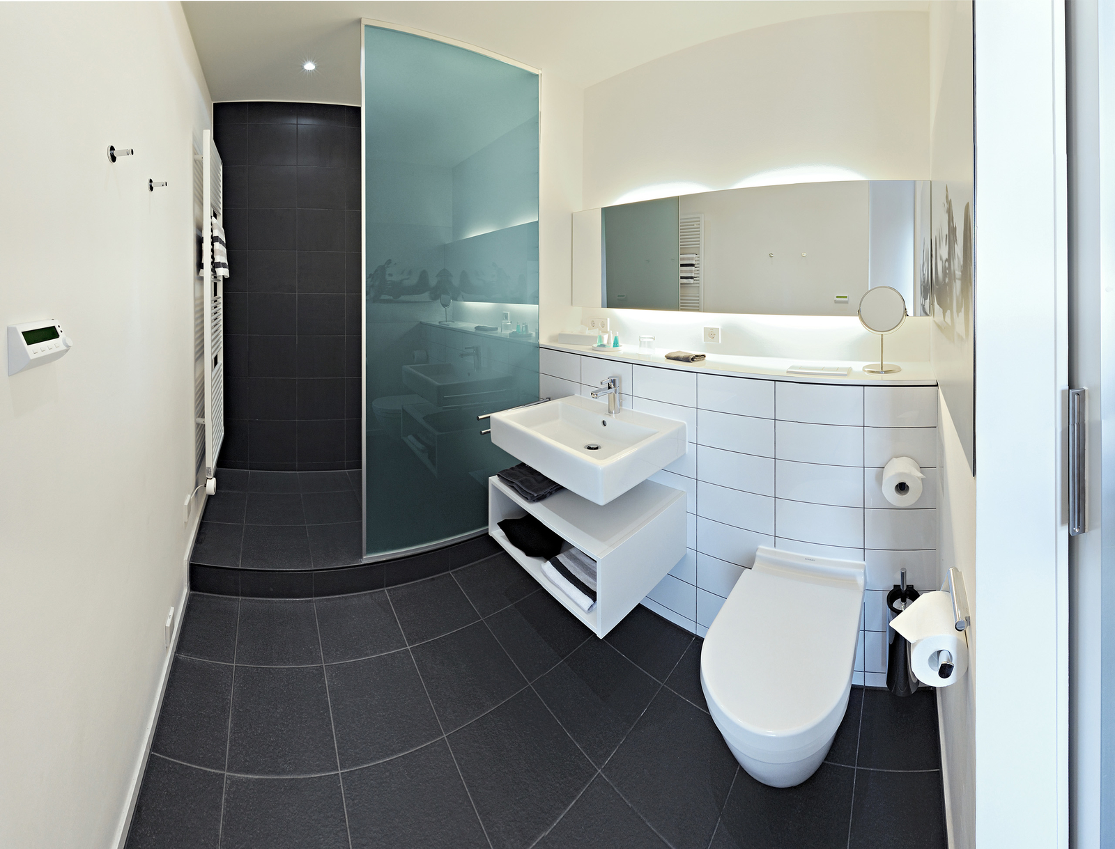 Ein modernes Badezimmer mit einer gläsernen Duschabtrennung, einem schwebenden Waschbecken und einem Wand-WC. Die Wände sind in neutralen Farbtönen gehalten, der Boden ist dunkel gefliest. Ein großer Spiegel sowie Handtücher und Toilettenpapier sind sichtbar.