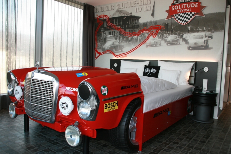 Ein rotes Bett in Form eines Mercedes-Rennwagens. Die Front des Fahrzeugs bildet die Kopfseite des Bettes, während die Matratze und Kissen im Hintergrund sichtbar sind. An der Wand im Hintergrund sind Rennstreckenbilder und nostalgische Autos abgebildet.