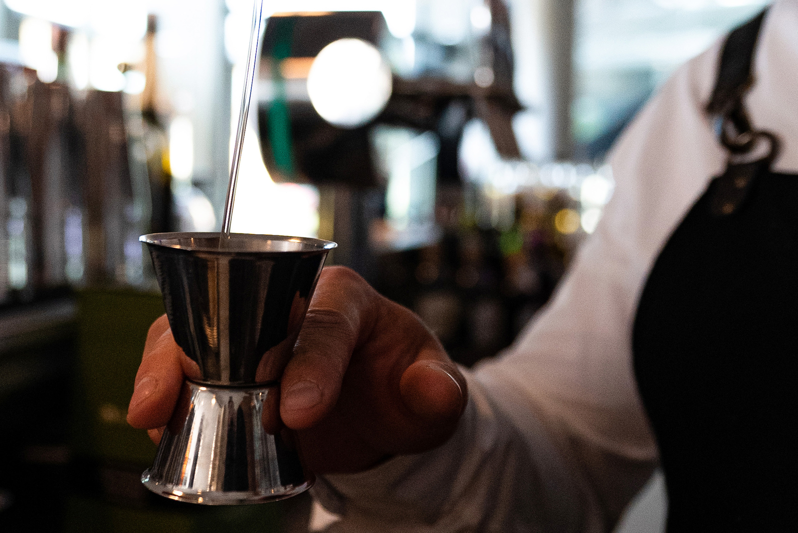 Ein Barkeeper hält einen Messbecher in der Hand, während er Flüssigkeit aus einem Ausgießer in den Becher gießt. Im Hintergrund sind verschwommen Flaschen und Barutensilien zu sehen. Die Atmosphäre wirkt lebhaft und geschäftig.