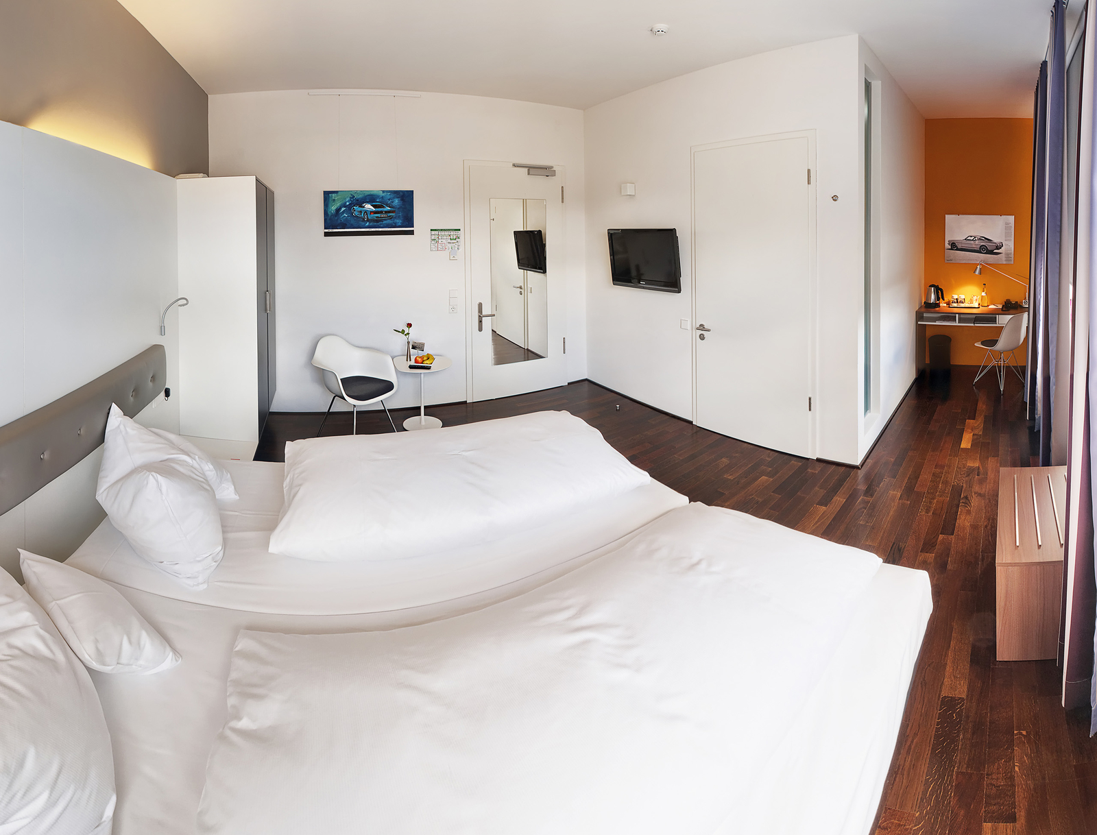 Ein helles, barrierefreies Hotelzimmer mit einem großen Doppelbett, einem Schreibtisch und einem Stuhl. An der Wand hängt ein Fernseher und ein Bild. Ein kleiner Tisch mit einer Blume und Früchten steht neben einem weißen Stuhl. Der Boden ist aus dunklem Holz.