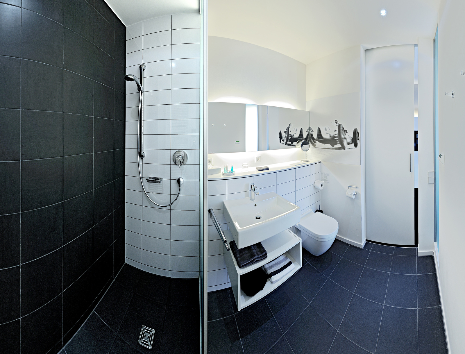 Das Bild zeigt ein modernes Badezimmer mit einer Dusche, einer Waschbecken und einem WC. Die Wände sind teilweise mit schwarzen und weißen Fliesen gestaltet. Ein Spiegel sowie Pflegeprodukte sind auf dem Waschtisch sichtbar. Die Einrichtung wirkt sauber und zeitgemäß.