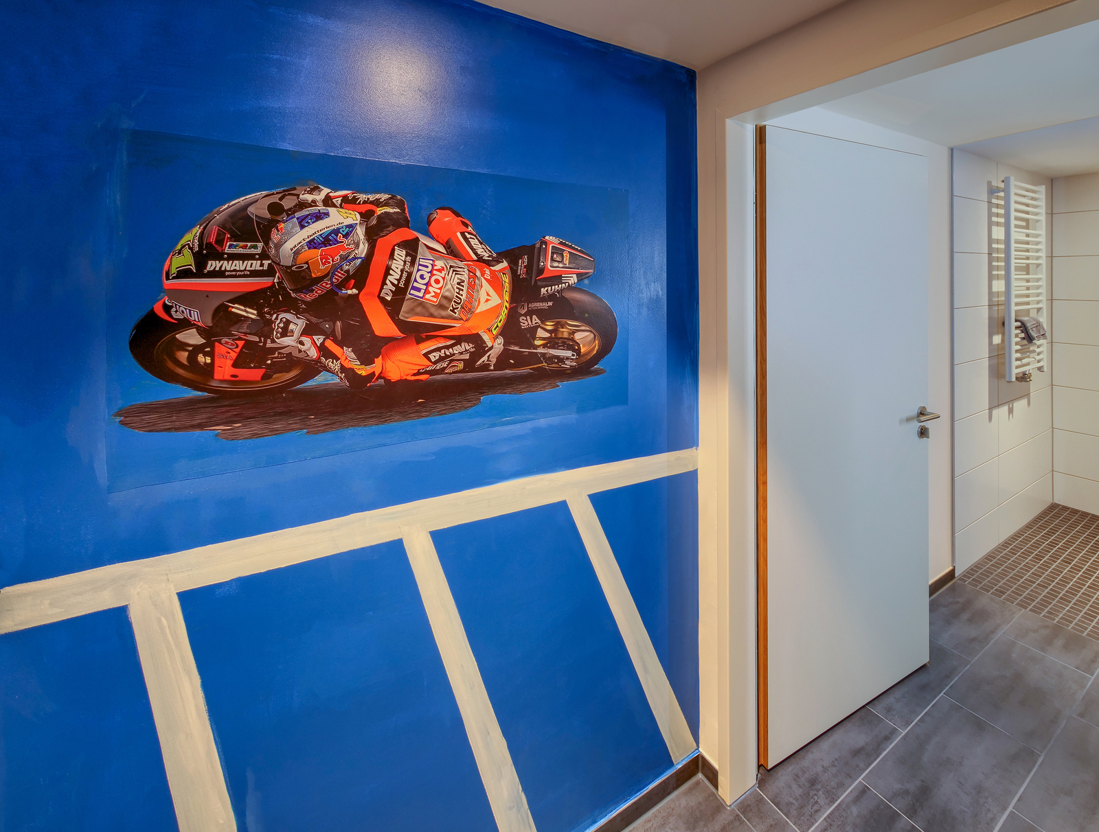 Eine Wandmalerei zeigt ein Motorrad, das schnell fährt, auf einem hellblauen Hintergrund. Der Fahrer trägt einen Helm und eine überwiegend schwarze und orangefarbene Rennkleidung. Die Bildkomposition vermittelt ein Gefühl von Geschwindigkeit und Dynamik.