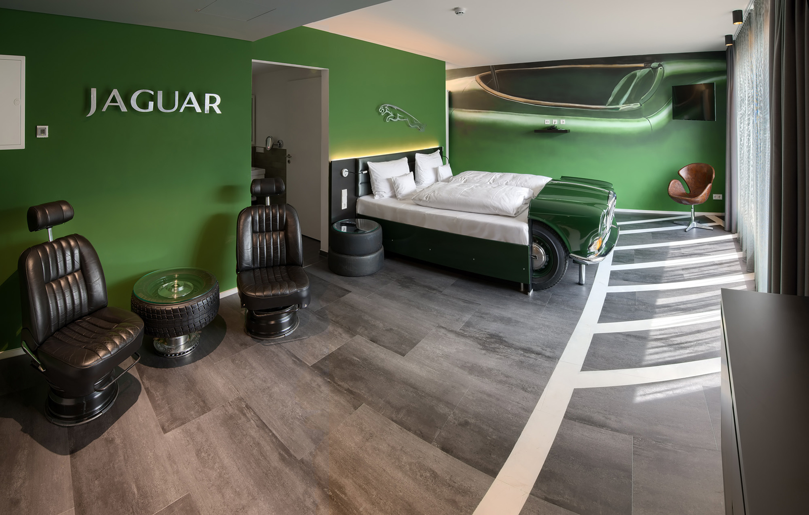 Das Bild zeigt ein modern gestaltetes Hotelzimmer mit grünen Wänden. Im Zentrum steht ein Bett, das wie ein Jaguar-Auto aussieht. Daneben befinden sich zwei schwarze Sessel und ein Tisch aus einem Autoreifen. Die Gesamtatmosphäre ist stilvoll und autobegeistert.