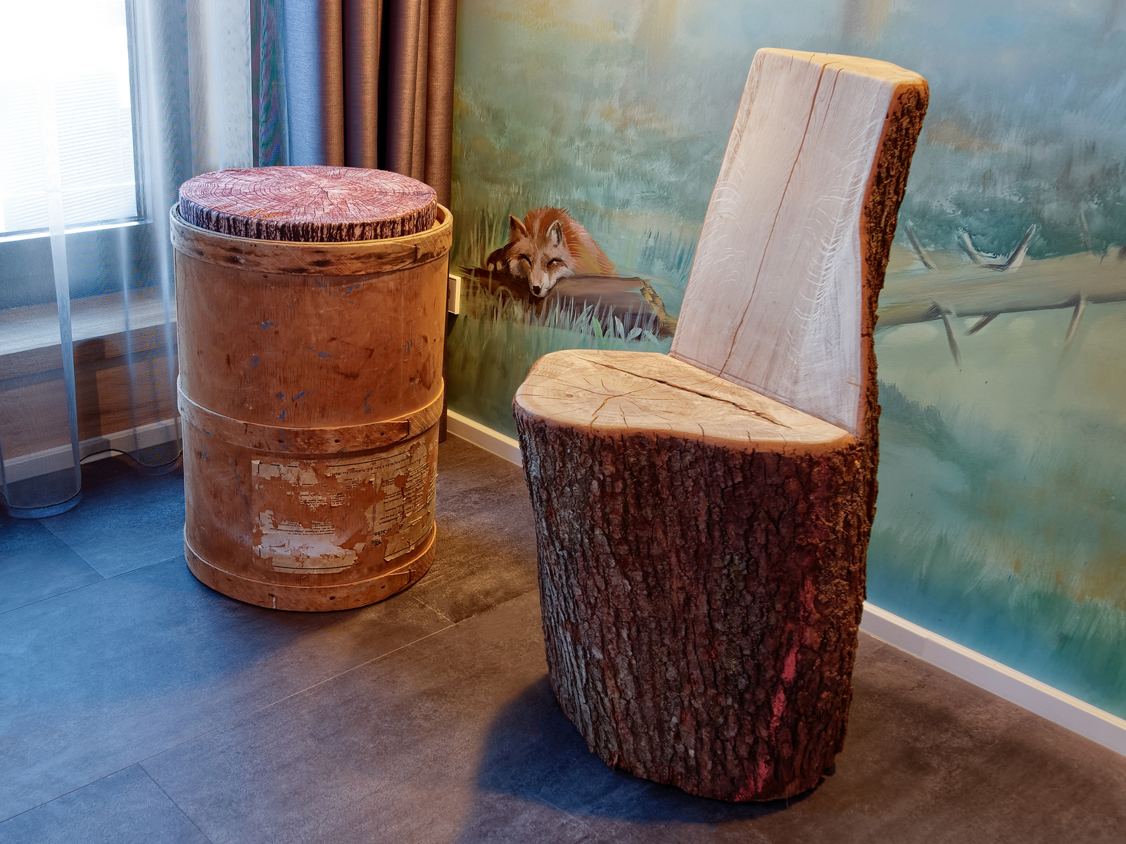 Ein rustikaler Holzstuhl in Baumstamm-Form steht neben einem runden, hölzernen Hocker mit einem gepolsterten Sitz. Die Möbel sind in einem Raum mit sanften Farben platziert, der einen natürlichen und gemütlichen Eindruck vermittelt.