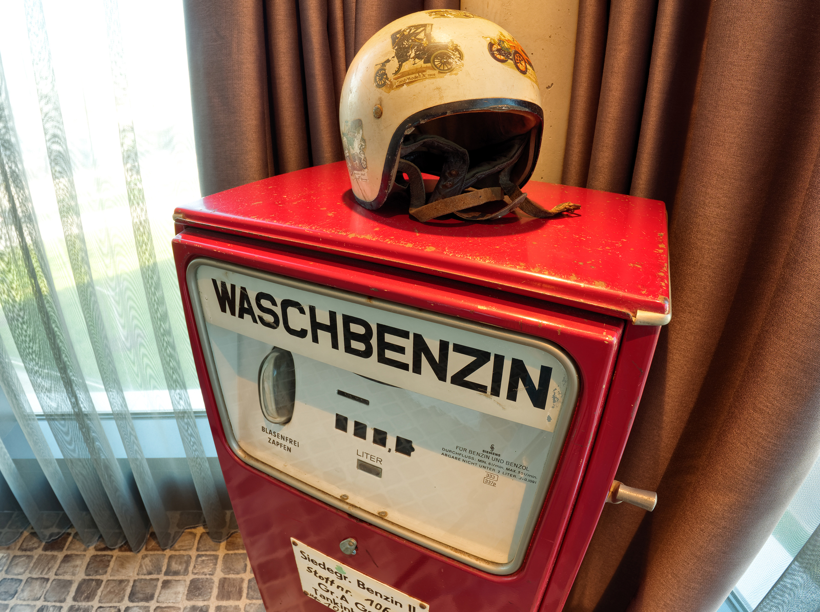 Ein roter Verkaufsautomat für Waschbenzin steht auf einem Tisch. Auf dem Automaten befindet sich ein Helm mit Aufklebern. Die Umgebung ist hell und modern, mit Vorhängen im Hintergrund.