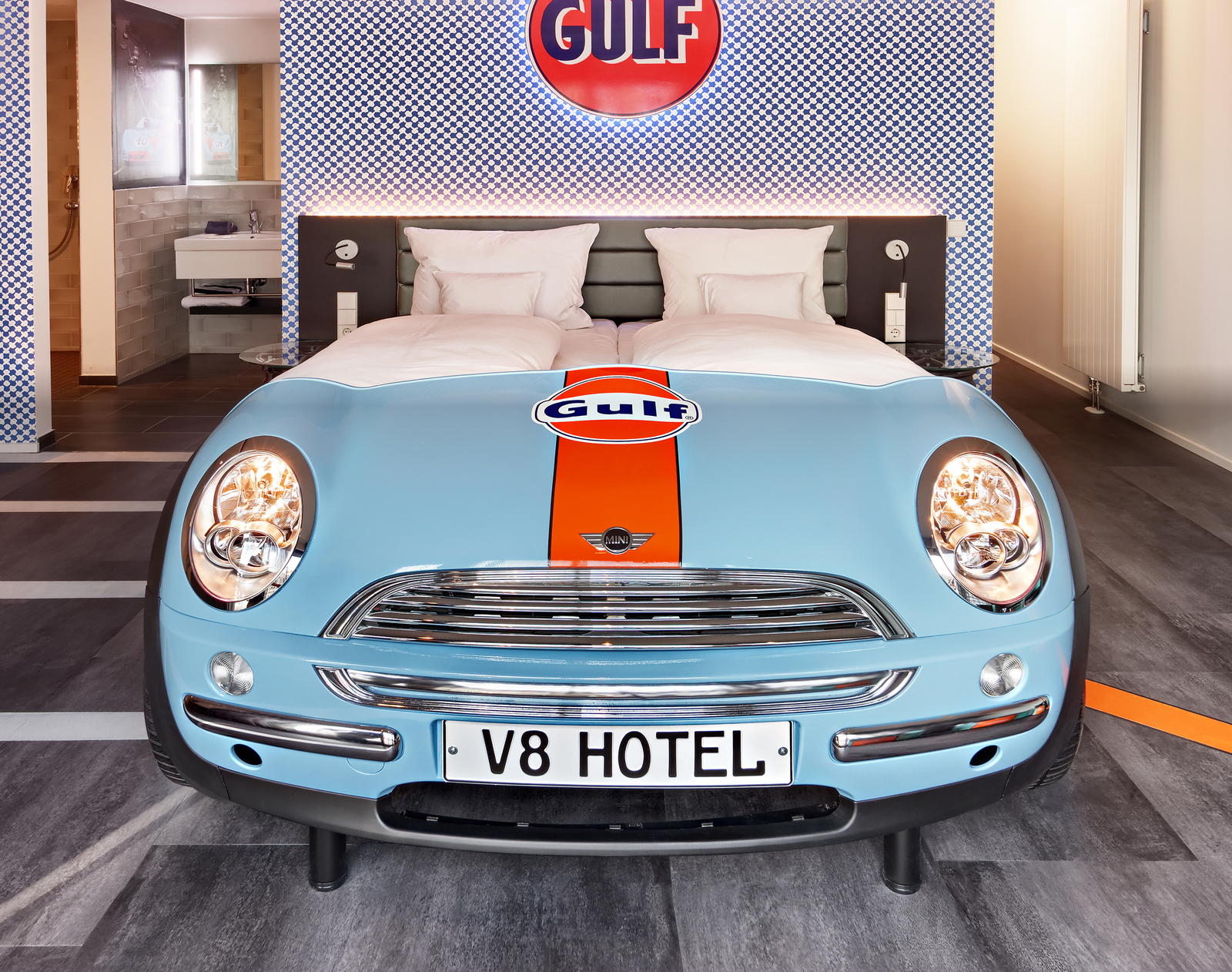 Hellblaues Autobett von Mini vor karierter Wand mit orangefarbenem Gulf-Logo im Autozimmer des V8 Hotels.
