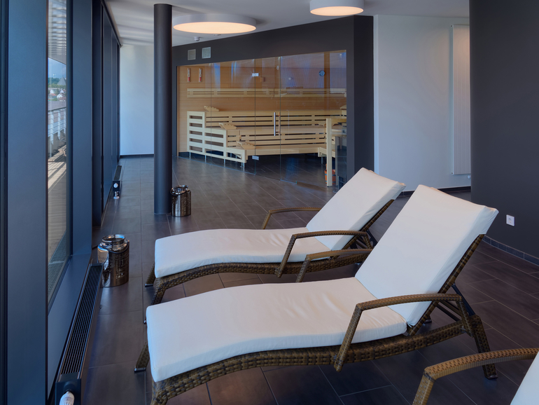 Blick auf zwei Liegestühle im Wellnessbereich des Hotels mit großer Holzsauna im Hintergrund.
