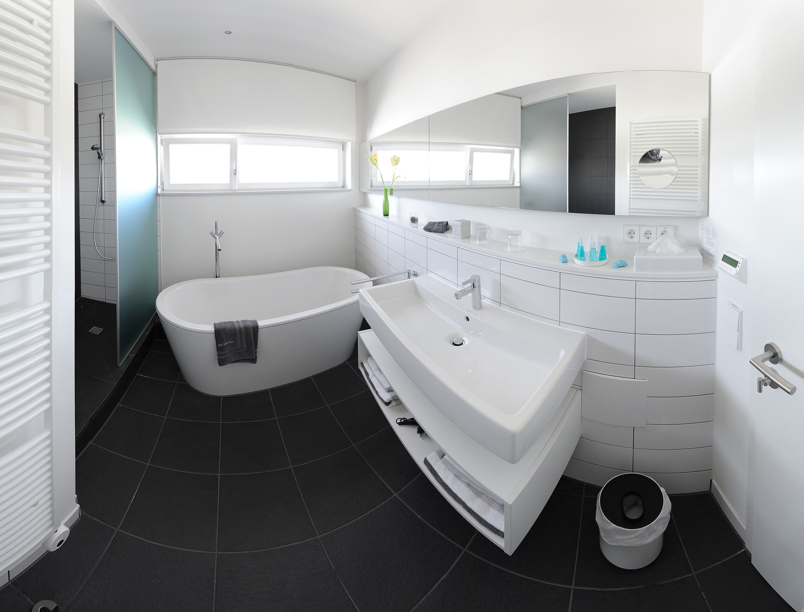 Das Bild zeigt ein modernes Badezimmer mit einer freistehenden Badewanne, einem Waschbecken mit Unterschrank und einer Dusche, die durch eine Glaswand abgetrennt ist. Eine kleine Pflanze steht auf dem Waschbecken. Der Boden ist mit dunklen Fliesen gestaltet.