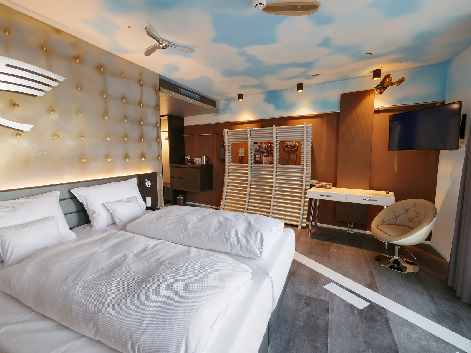 Ein modernes Hotelzimmer mit zwei Einzelbetten, weißen Bettdecken und Kopfkissen. An der Wand befindet sich eine dekorative, gepolsterte Fläche. Im Hintergrund ist ein Fernseher, ein Tisch und ein Stuhl sichtbar. Die Decke ist in sanften Himmelstönen gestaltet.
