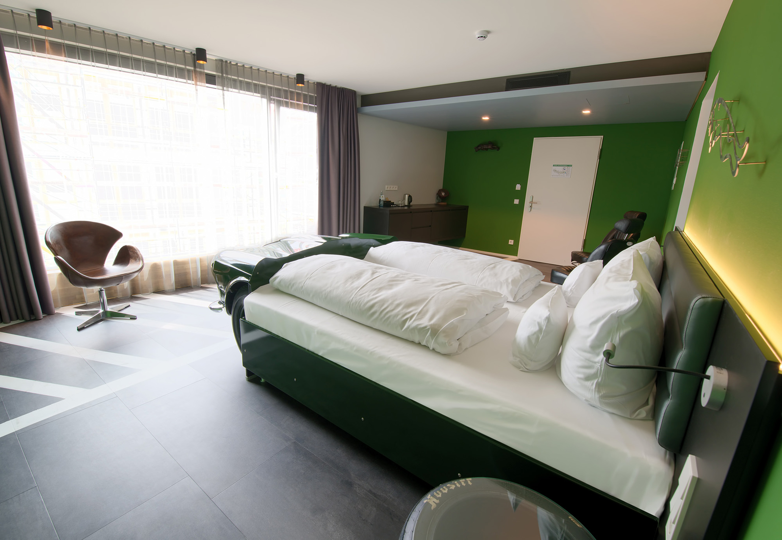 Das Bild zeigt ein modernes Hotelzimmer mit einem großen Doppelbett, das mit weißen Kissen ausgestattet ist. An der Wand befindet sich eine Grünfläche. In der Ecke steht ein stylischer Sessel. Der Raum hat große Fenster, die viel Tageslicht hereinlassen.