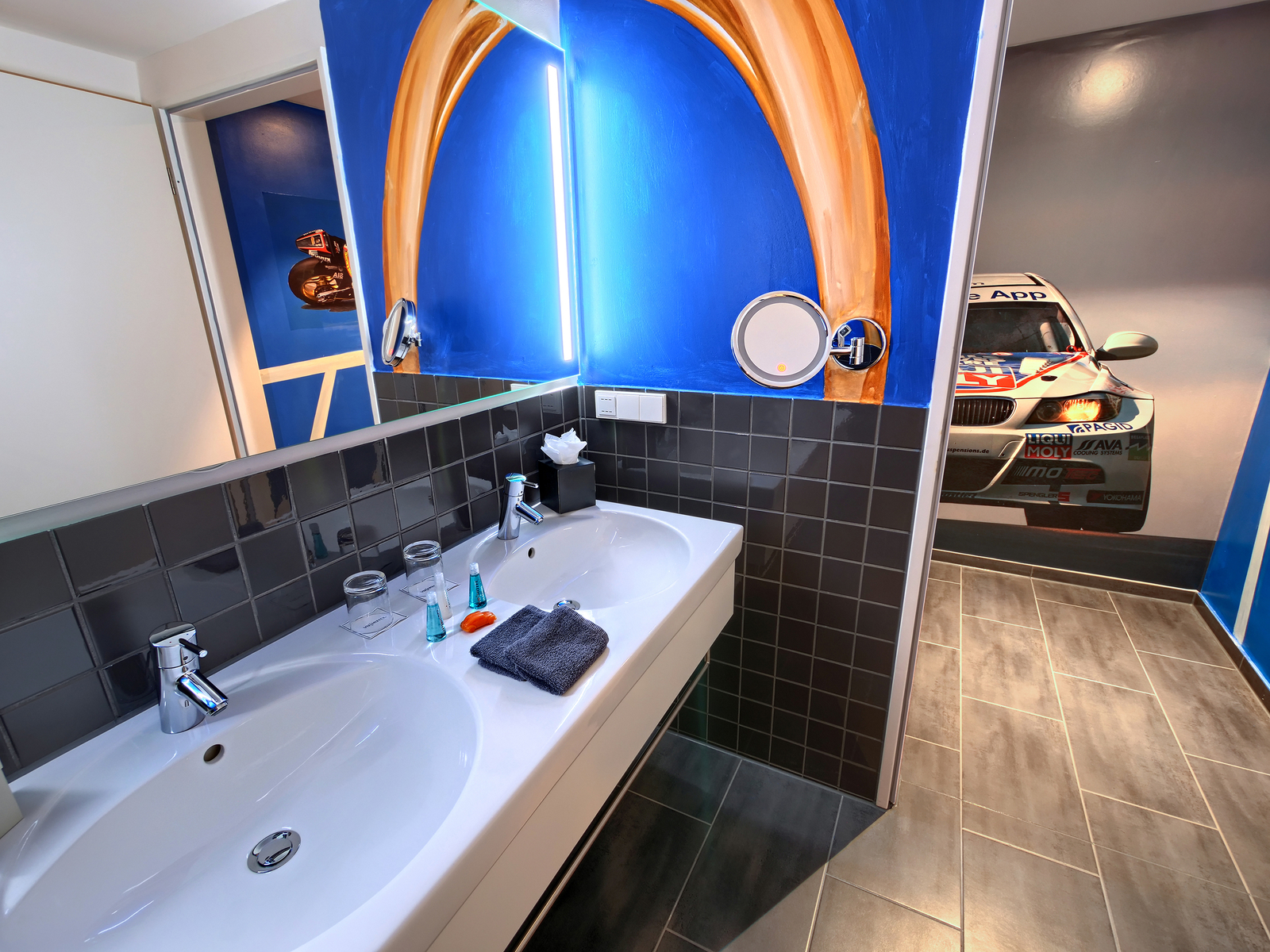 Ein modernes Badezimmer mit doppeltem Waschbecken, eleganten Wasserhähnen und einer Spiegelbeleuchtung. Der Boden ist mit grauen Fliesen ausgelegt, während die Wände in lebendigen Blau- und Grautönen gestaltet sind. Eine Tür führt zu einem Bereich mit einem überdimensionalen Auto-Motiv.