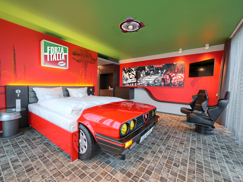 Autozimmer Forza Italia mit rotem Autobett neben einer Fensterfront, rotem Stuhl und roter Zapfsäule.
