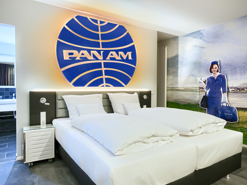Das Bild zeigt ein modernes Hotelzimmer mit einem großen PAN AM-Logo an der Wand. Im Hintergrund steht eine Figur einer Stewardess in einem blauen Uniformanzug mit einer Tasche. Das Zimmer ist hell gestaltet, mit einem großen Doppelbett und eleganten Möbeln.