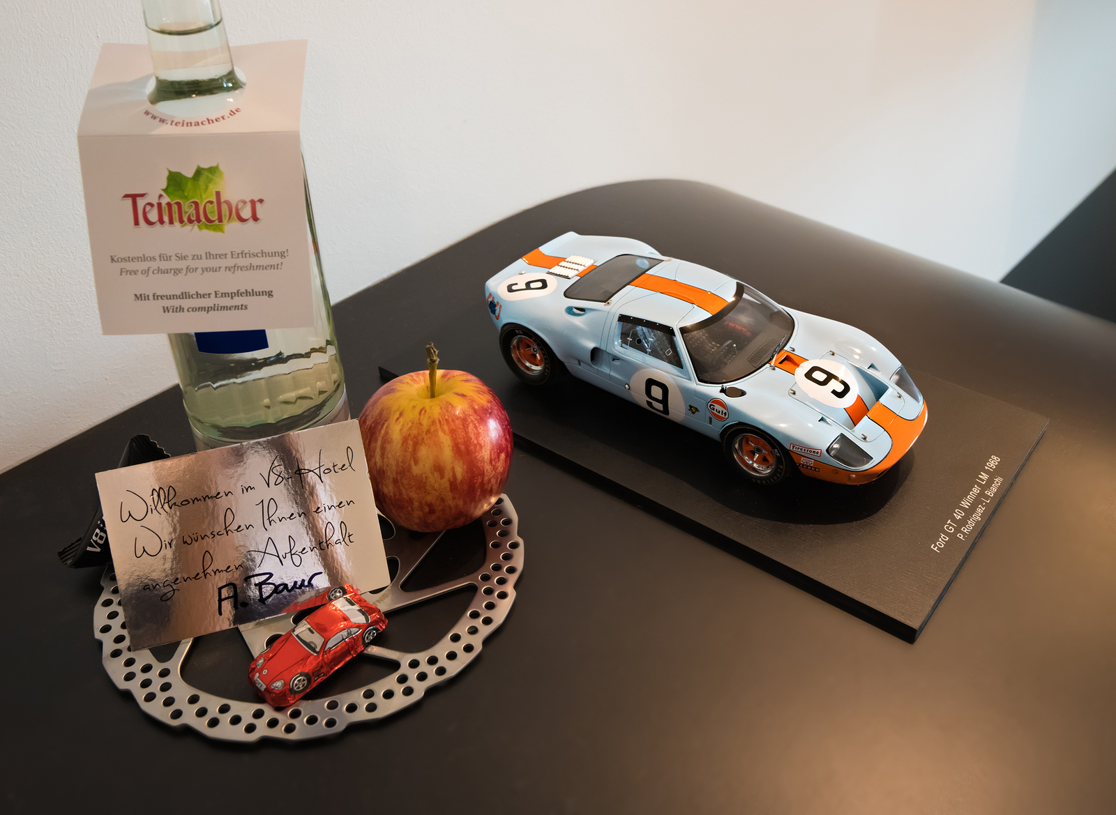 Ein dekorativer Tisch mit einer Flasche Wasser, einem roten Apfel und einer kleinen, roten Spielzeugautos. Auf einem Notizblatt steht eine Willkommensnachricht für Gäste. Daneben ist ein Modell eines blauen Sportwagens mit orangefarbenen Streifen.