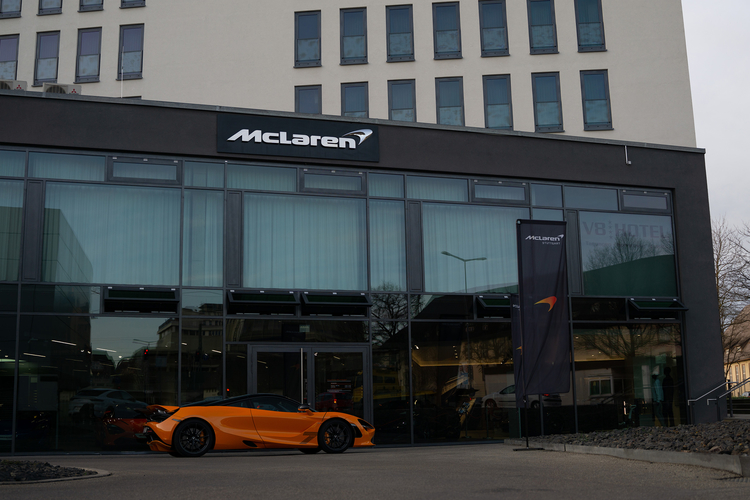 Ein orangefarbener Sportwagen steht vor einem modernen Gebäude mit großen Glasfenstern. Über dem Eingang prangt das McLaren-Logo. Im Hintergrund sind weitere Fahrzeuge und eine Fahne mit dem McLaren-Design sichtbar. Das Gebäude klingt nach einem Verkaufsstandort oder einer Ausstellung für Luxusautos.
